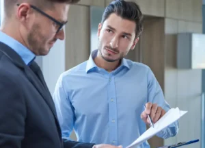 An auditor and a manager engage in a pre-assessment review, going over quality or compliance documentation using a clipboard and tablet in a professional office setting