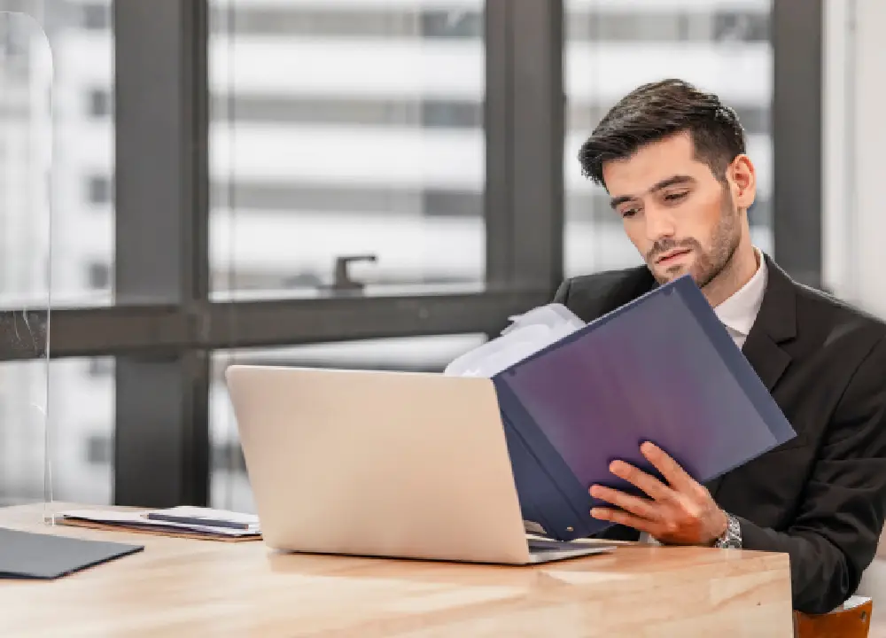 Professional reviewing quality compliance documents on a laptop and binder